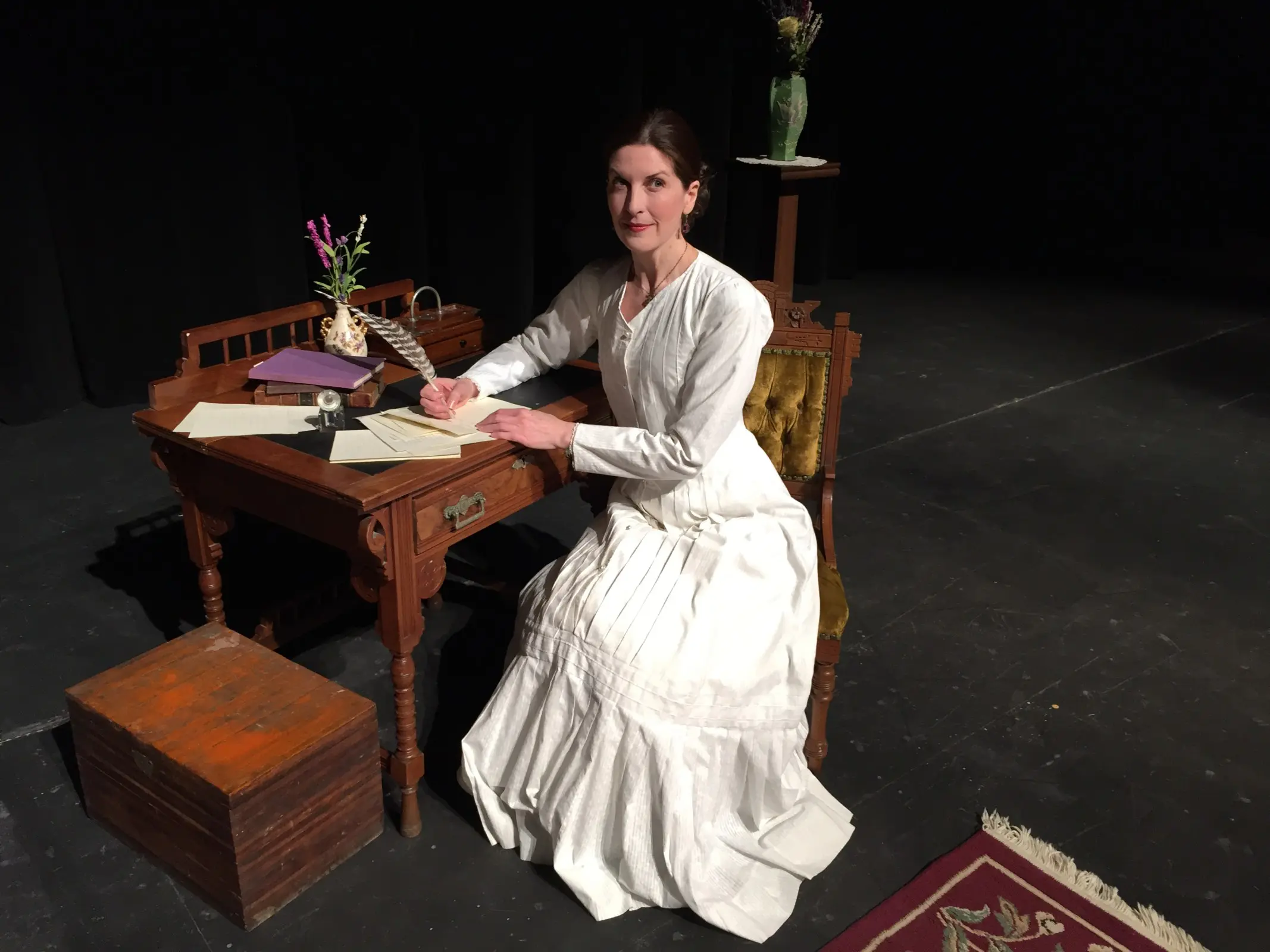 Period costume woman seated at wooden desk.