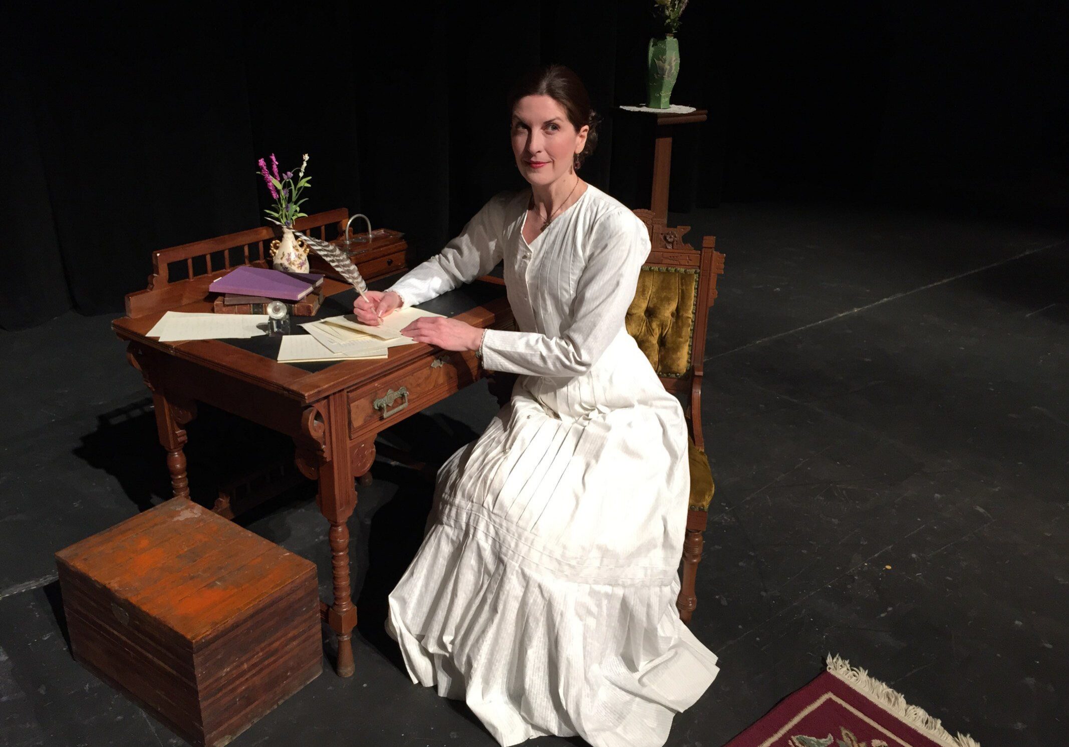 Period costume woman seated at wooden desk.